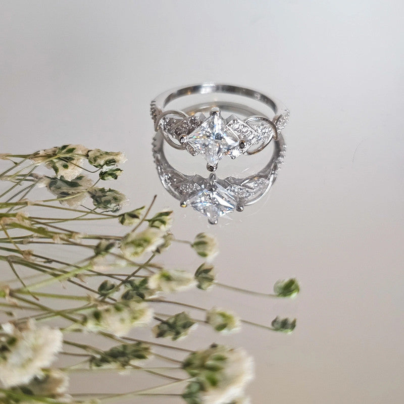 Moissanite sterling silver ring with accent stones reflected on light surface near delicate dried flowers
