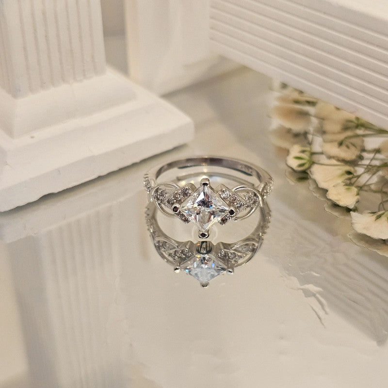 Moissanite sterling silver ring with square-cut center stone and accent stones reflected on surface, framed by white columns and dried flowers