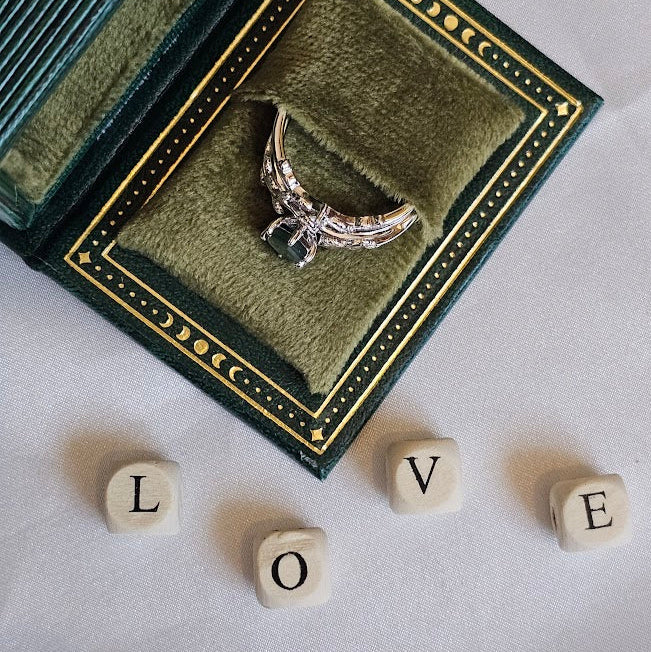 Silver ring with wings in a green jewelry box on a white surface with 'LOVE' spelled out with dice.