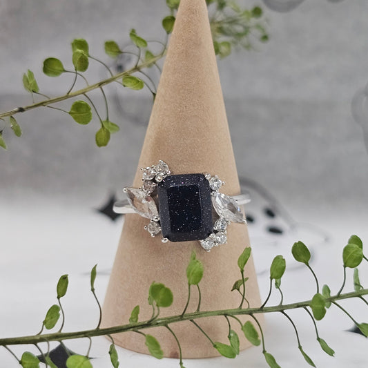 Black square stone ring on a wooden stand with green leaves in the background