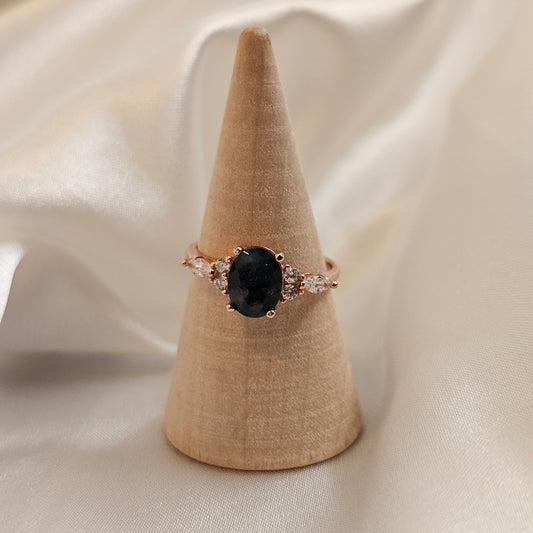 Ring with a dark green moss agate gemstone and rose gold accents on a beige stand against a beige fabric background