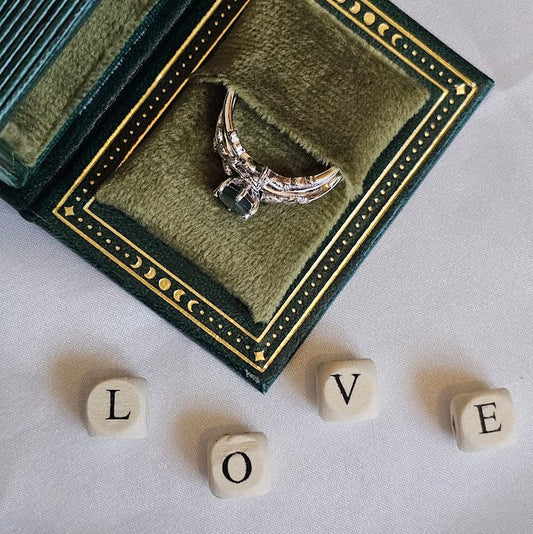 Silver ring with wings in a green jewelry box on a white surface with 'LOVE' spelled out with dice.