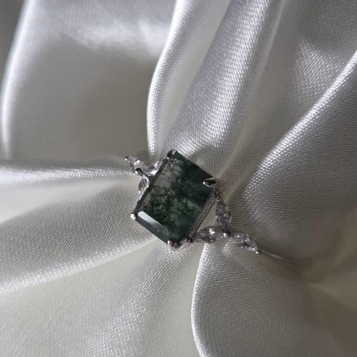 Natural moss agate ring with a square-cut moss agate centre stone featuring rich green moss-like inclusions, set in solid 925 sterling silver with delicate white cubic zirconia accents. Styled against soft white satin, this nature-inspired moss agate ring highlights the organic woodland patterns within the gemstone and the elegant silver band design, part of the Whispers of the Forest collection by Moonstone & Sage.