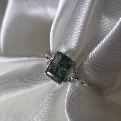 Natural moss agate ring with a square-cut moss agate centre stone featuring rich green moss-like inclusions, set in solid 925 sterling silver with delicate white cubic zirconia accents. Styled against soft white satin, this nature-inspired moss agate ring highlights the organic woodland patterns within the gemstone and the elegant silver band design, part of the Whispers of the Forest collection by Moonstone & Sage.