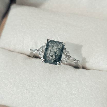 Close-up of a natural moss agate ring featuring a square-cut moss agate centre stone with distinctive dark green moss-like inclusions, set in solid 925 sterling silver. The moss agate gemstone is framed by delicate white cubic zirconia accents on either side, highlighting the organic patterns within the stone. This nature-inspired moss agate ring showcases a unique one-of-a-kind gemstone, part of the Whispers of the Forest collection by Moonstone & Sage.