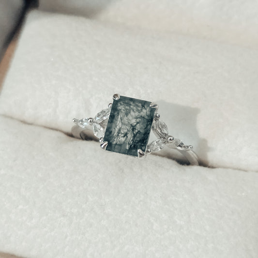 Close-up of a natural moss agate ring featuring a square-cut moss agate centre stone with distinctive dark green moss-like inclusions, set in solid 925 sterling silver. The moss agate gemstone is framed by delicate white cubic zirconia accents on either side, highlighting the organic patterns within the stone. This nature-inspired moss agate ring showcases a unique one-of-a-kind gemstone, part of the Whispers of the Forest collection by Moonstone & Sage.