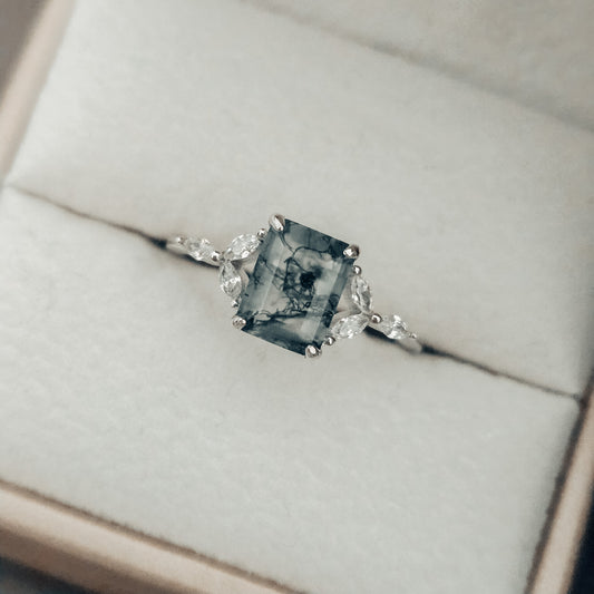 Close-up of a natural moss agate ring featuring a square-cut moss agate centre stone with distinctive dark green moss-like inclusions, set in solid 925 sterling silver. The moss agate gemstone is framed by delicate white cubic zirconia accents on either side, highlighting the organic patterns within the stone. This nature-inspired moss agate ring showcases a unique one-of-a-kind gemstone, part of the Whispers of the Forest collection by Moonstone & Sage.