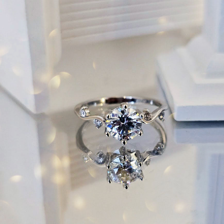 Moissanite engagement ring with round brilliant-cut center stone and twisting vine band with CZ accents displayed on reflective white surface