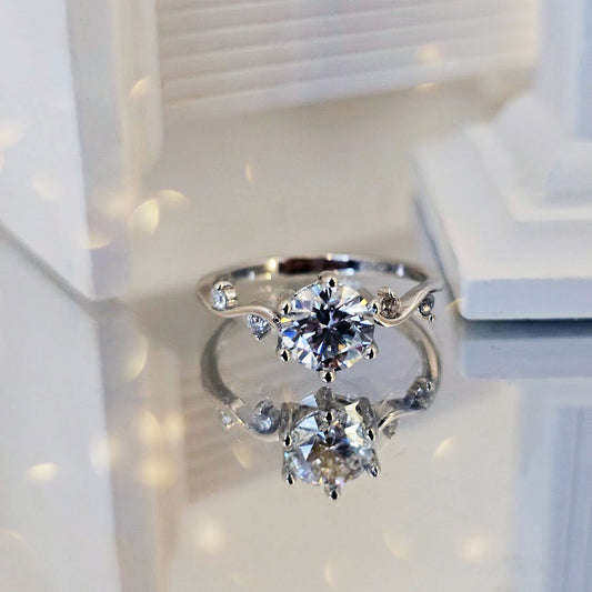 Moissanite engagement ring with round brilliant-cut center stone and twisting vine band with CZ accents displayed on reflective white surface
