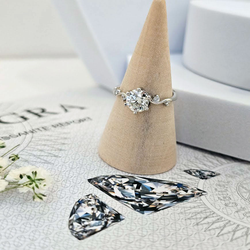 Moissanite engagement ring with twisting vine band and round brilliant-cut center stone displayed on beige cone stand on GRA certificate paper with diamond illustrations