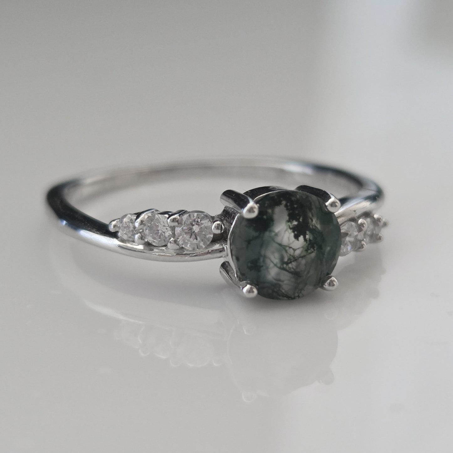 Close-up of a natural moss agate ring featuring a round moss agate gemstone with delicate cubic zirconia accent stones, crafted in 925 sterling silver and photographed on a soft white background.