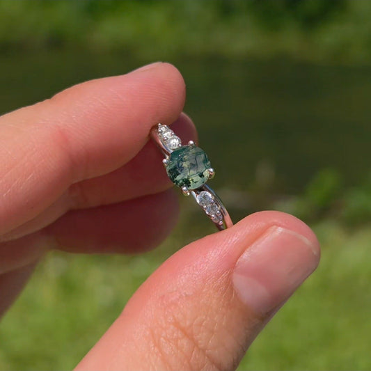 Hand holding a natural moss agate ring with an oval green moss agate gemstone and cubic zirconia accents in 925 sterling silver against a soft green outdoor background.