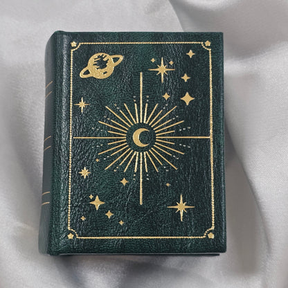 Green book style ring box with gold celestial designs on a white fabric background