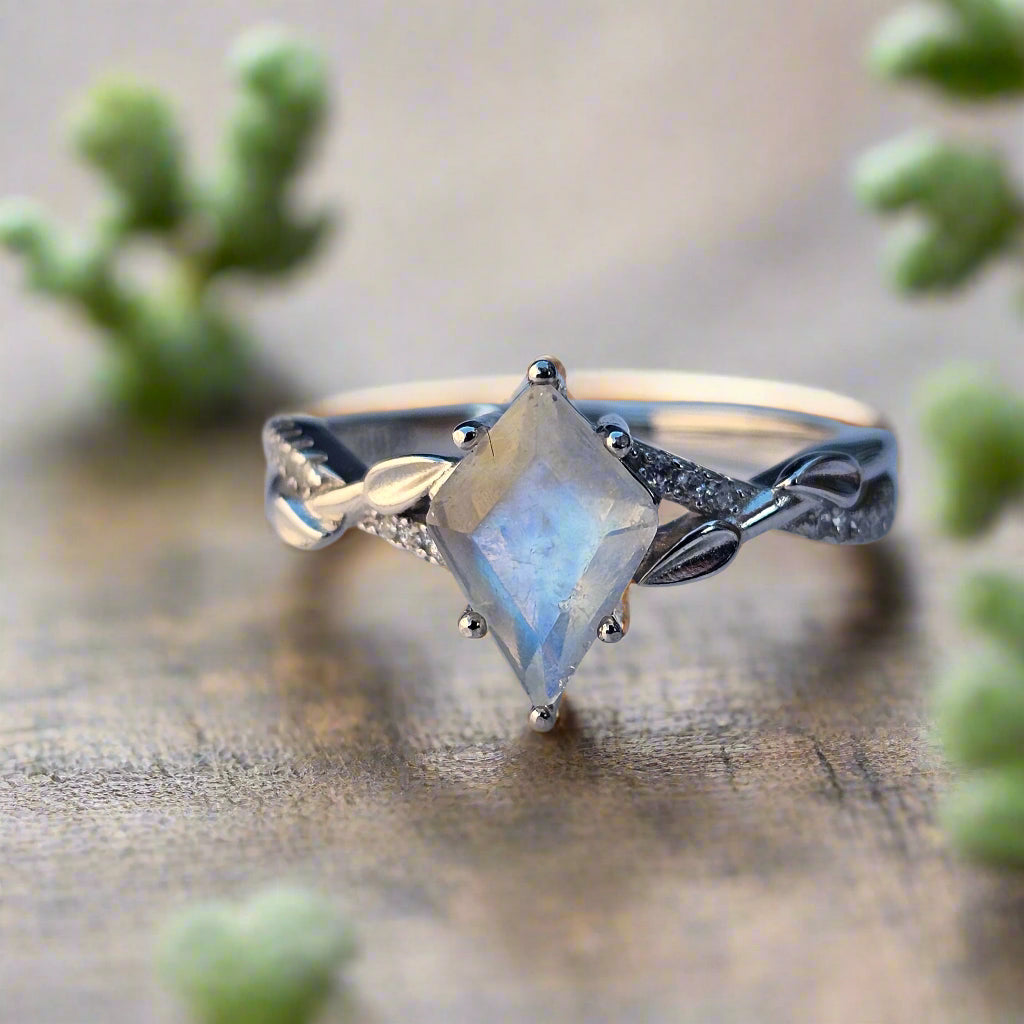 Mystic Moonstone Ring in 925 sterling silver with 18K white gold plating, featuring a kite-cut natural moonstone and cubic zirconia accent stones, displayed on a nature-inspired wooden surface with soft green foliage.